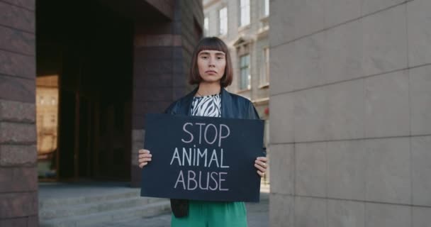 Молодая женщина с кольцом в носу держит плакат с остановкой жестокого обращения с животными, стоя на городской улице. Тысячелетняя девочка бьет против жестокости. Концепция социальных проблем. Увеличь. — стоковое видео