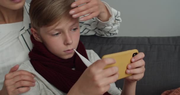 Close up widok chorego dziecka gry online na smartfonie i pomiaru temperatury.Mother dotykając jej syna czoło i biorąc termometr, gdy siedzą na kanapie w domu. — Wideo stockowe