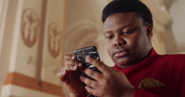 Nahaufnahme eines Afroamerikaners im roten Anzug mit seinem Smartphone. Junge männliche Person, die auf der Suche nach Gospelliedern den Bildschirm des Telefons anschaut und berührt. Innenräume. — Stockvideo