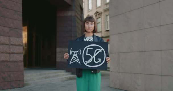 Jeune femme hipster tenant une pancarte sans signe de 5g. Fille millénaire protestant contre la technologie 5G et le déploiement d'antenne compatible 5G tout en se tenant debout dans la rue. Zoom avant. — Video