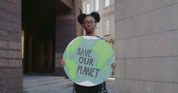 Chica afroamericana con cartel de cartón mirando a la cámara mientras apoya el movimiento ecológico. Mujer milenaria sosteniendo modelo de tierra con salvar nuestro planeta escribiendo mientras está de pie en la calle. — Vídeo de stock
