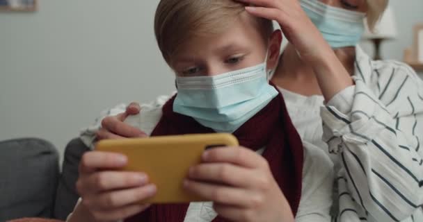 Close up view of woman and her teen son wearing medical masks and looking at phone screen. Teen boy playing online game on smartphone while sitting with mom on sofa at home. — Stock Video
