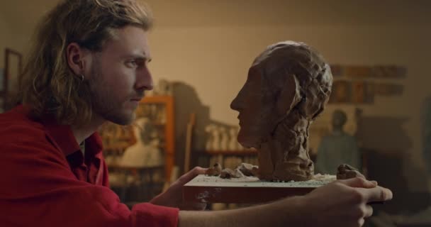 Talentueux jeune homme tournant la tête de sculpture de l'argile de poterie qui se tient debout sur le postament. Beau sculpteur regardant sa création inachevée tout en étant dans l'atelier de sculpture. — Video