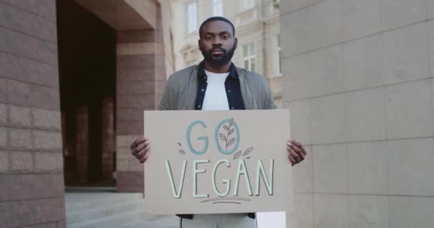Portret afrykańskiego faceta trzymającego Go Vegan sztandar stojąc na ulicy.Przystojny aktywista męski agitujący wegitariański styl życia. Koncepcja go zielony i weganizm. — Wideo stockowe