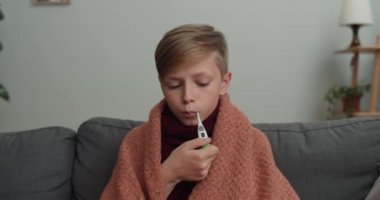 Çocuk kanepede otururken üşümüş ve vücut ısısını ölçüyormuş. Genç çocuk battaniyeye sarılı termometreye bakıyor ve başını sallıyor. Hastalık ve sağlık sigortası kavramı.