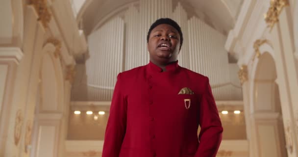 Homem afro-americano a cantar música gospel. Jovem cantor em terno vermelho realizando emocionalmente e mãos em movimento enquanto está de pé na igreja. Conceito de pessoas e religião. — Vídeo de Stock