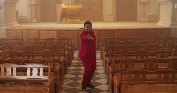 Afro american young musician wearing in stylish red suit dancing while singing song. Male cheerful performer of gospel music moving body while standing at row of wooden pews. Indoors. — Stock Video