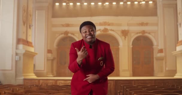 Joven músico masculino con ropa roja mirando a la cámara y cantando mientras está parado en la fila de bancos de madera. Chico alegre interpretando música gospel y moviendo las manos. En interiores. — Vídeo de stock