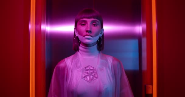 Portrait of trendy female clubber turning head and looking to camera. Young woman with make up wearing fashionable clothes posing to camera while standing in room with neon lights. — Stock Video