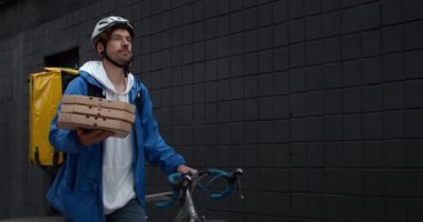 Bisikletli, koruyucu kasklı, çantalı ve bisikletli genç yakışıklı adam sokakta düzeni ve bisikleti taşıyordu. Bisikletli erkek kurye müşteriye pizza getiriyor. Yemek servisi kavramı.