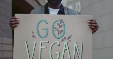 Yakışıklı erkek aktivistin sokakta dikilirken elinde vegan kartonu tutmasını yakından izleyin. Sakallı Afro-Amerikan adam vejetaryenliği ve sağlıklı yaşam tarzını destekliyor..
