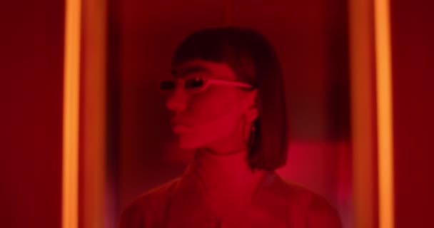 Portræt af ung stilfuld kvinde i futuristiske briller dreje hovedet og ser til kameraet. Nærbillede af kvindelig klubber med næsering poserer på neon baggrund. Zoom ind. – Stock-video