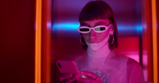 Vedere de aproape a unei tinere femei în ochelari de soare la modă care navighează pe internet în timp ce stă în cameră cu lumină de neon. Femeie cu machiaj futurist folosind smartphone-ul și uitându-se la ecran . — Videoclip de stoc