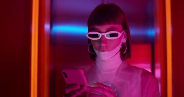 Vedere culturală a femeii la modă în ochelari de soare moderni defilare ecran în timp ce stați în cameră cu lumini de neon. Modelul feminin cu machiaj futurist folosind telefonul mobil și uitându-se la ecran . — Videoclip de stoc