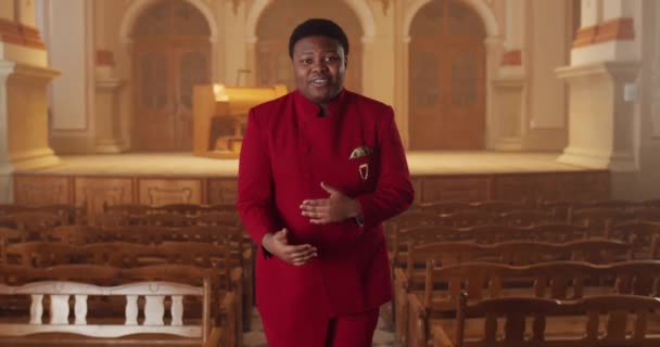 Afro american male performer in red suit looking to camera and singing while standing at row of wooden pews in hall. Smiling guy performing gospel music emotionally and dancing. — Stock Video