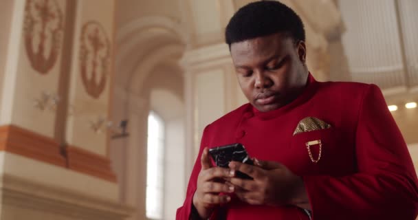 Portret van Afro-Amerikaanse man in rood pak met zijn smartphone. Jonge mannelijke persoon op zoek en aanraken van het telefoonscherm tijdens het zoeken gospel song woorden. Binnenshuis. — Stockvideo