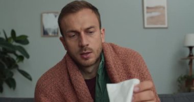 Genç adamın mendil alıp rahat bir odada kanepede otururken hapşırmasını yakından izleyin. Sakallı adam hastalanıyor ve evde vakit geçiriyor. Hastalık ve grip kavramı.