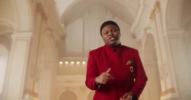 Bottom view of gospel singer in red suit looking to camera and singing while standing at church hall. Young guy clapping and moving hands in rhytm while performing worship music. — Stock Video