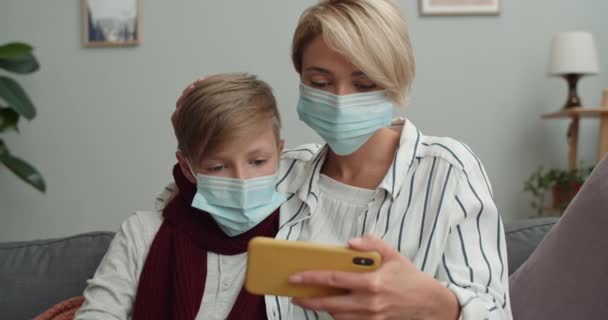 Vista Crop di famiglia malata utilizzando telefono whilw seduto sul divano a casa. Donna e suo figlio adolescente in maschere mediche guardando lo schermo dello smartphone durante la visione di video. Concetto di malattia. — Video Stock