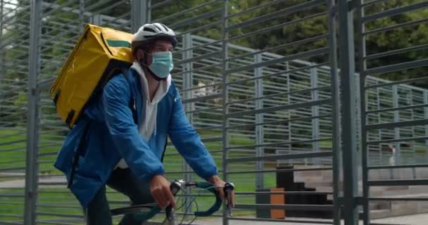 Junger Fahrradkurier mit medizinischer Maske auf dem Gesicht trägt einen Lebensmittelrucksack, während er auf der Straße unterwegs ist. Nahaufnahme eines Hipster-Mannes mit Schutzhelm, der dem Kunden Befehl gibt. — Stockvideo