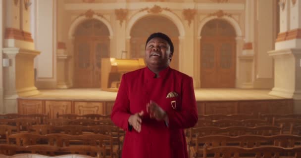 Cheerful african american gospel singer standing at row of wooden church pews and singing. Young man performing worship music and moving hands in rhytm.Concept of people and religion. — Stock Video