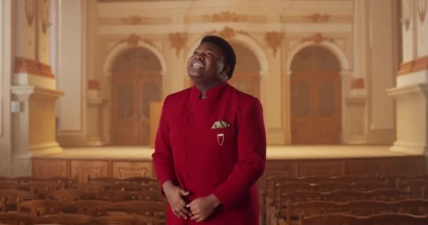 Musicien masculin talentueux en costume rouge chantant tout en se tenant à la rangée de bancs en bois dans le hall. Joyfull guy jouer de la musique gospel émotionnellement et bouger les mains. À l'intérieur. — Video