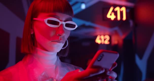 Attractive woman in futuristic shades typing message while communicating in social media. Female clubber with make up using her smartphone while standing in corridor with neon lighting. — Stock Video