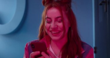 Neon ışıklı odada otururken akıllı telefon kullanan güzel bir genç kadının görüntüsünü kapat. Şık saç stili olan neşeli bayan kameraya bakıyor ve gülümsüyor..