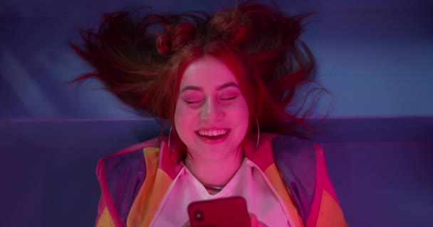 Bovenaanzicht van vrolijke aantrekkelijke vrouw met smartphone terwijl ze in de kamer ligt met neon verlichting. Millennial vrouwelijke persoon in trendy kleding lachen tijdens het chatten in sociale media. — Stockvideo