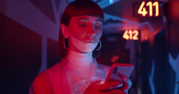 Close up de mulher usando telefone enquanto estava em pé no hall do hotel futurista com luz de néon e números brilhantes. Pessoa feminina com maquiagem olhando para a tela do smartphone. — Vídeo de Stock