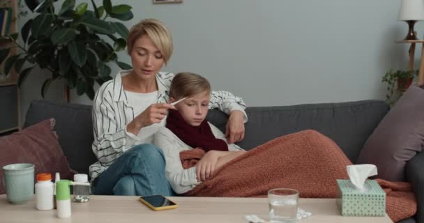 La mamma prende e guarda il termometro mentre è seduta sul divano con il bambino malato. Donna preoccupata chiama il medico sullo smartphone mentre suo figlio adolescente ha la febbre. Concetto di malattia. — Video Stock