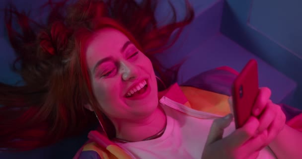 Vista lateral da jovem mulher feliz usando smartphone enquanto deitado em escadas com iluminação de néon. Boa aparência feminina em roupas da moda sorrindo enquanto conversa nas mídias sociais. — Vídeo de Stock