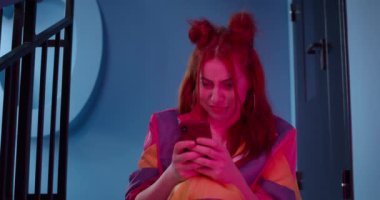 Telefon kullanırken gülümseyen şık elbiseli neşeli genç insan manzarası. Büyük küpeli bir kadın daktilo ediyor ve cep telefonu ekranına bakıyor. Neon ışıklandırma arkaplanı.