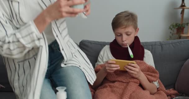 Schöne Mutter bringt ihrem Sohn Heißgetränk und nimmt Thermometer. Krankes Kind misst Temperatur und spielt auf dem Smartphone, während es auf dem Sofa sitzt. Krankheitsbegriff. — Stockvideo