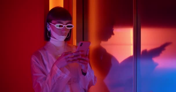 Vedere culturală a tânărului club feminin în ochelari de soare futuriști tastând și derulând în timp ce stătea lângă lift. Femeia cu machiaj poartă haine la modă folosind telefonul mobil . — Videoclip de stoc