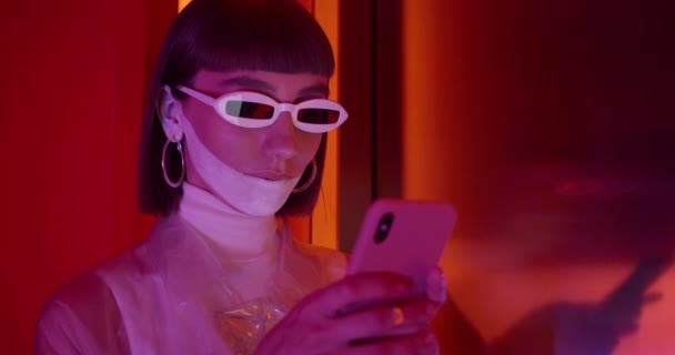 Close-up zicht op jonge vrouw in trendy zonnebril typen en scrollen terwijl staan in de kamer met neon licht. Vrouwelijke clubber met futuristische make-up en neusring met smartphone. — Stockvideo