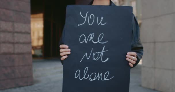 Female hands holding carton placard with you are not alone writing on it at city street. Concept of social problem and support. — Stock Video