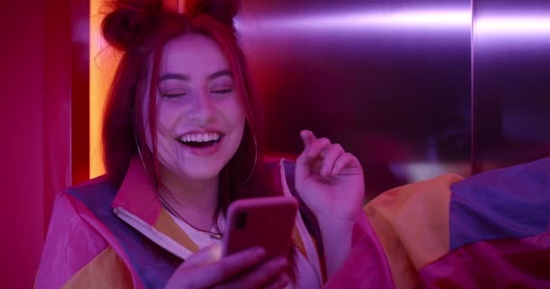 Widok na zboże młodej kobiety w modnym stroju i fryzury za pomocą smartfona i śmiechu. Szczęśliwego tysiąclecia kobieta osoba przewijania i patrząc na ekran telefonu siedząc w neonach. — Wideo stockowe