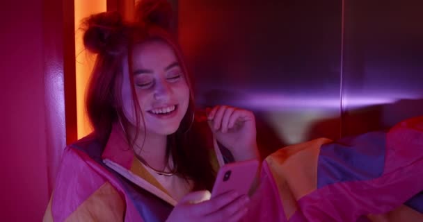 Millennial vrouw in modieuze kleding en kapsel scrollen neews voeden en glimlachen. Vrolijke vrouwelijke clubber met behulp van smartphone en kijken naar het scherm terwijl zitten in neon lichten. — Stockvideo