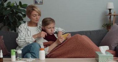 Genç çocuk sıcaklığı ölçmek için battaniyeyi örttü ve akıllı telefondan annesine yaslanırken oyun oynadı. Güzel bir kadın ve hasta oğlu evdeki kanepede oturuyor..
