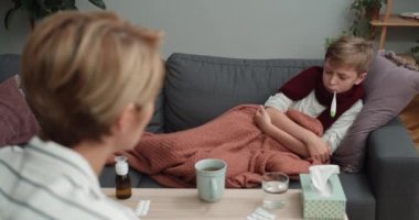 Tenisçi çocuk sıcaklığı ölçerken annesini dinliyor. Arka planda hasta oğluyla konuşan bir kadın var. O, evdeki kanepede yatıyor. Sağlık ve hastalık kavramı.