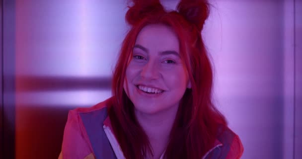 Portret radosnej młodej kobiety odwracającej głowę i patrzącej w kamerę. Close up widok tysiąclecia kobieta clubber w modnym stroju i fryzury possing siedząc w neon oświetlenia. — Wideo stockowe