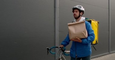 Sırt çantalı, sokakta bisiklet iten erkek teslimatçının ön görüntüsü. Siyah duvarın yanında yürürken yanında kağıt paket taşıyan koruyucu kasklı bir adam..