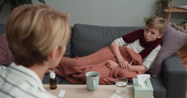 Hasta oğluyla evdeki kanepede yatarken iletişim kuran kadının arka plandaki görüntüsü. Çocuk annesiyle konuşuyor ve termometreyi tutarken gülümsüyor. Sağlık ve hastalık kavramı. — Stok video