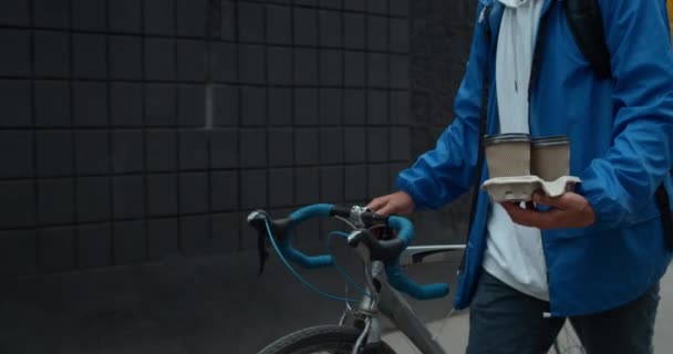 Widok na zboże męskiego kuriera niosącego kubki kartonowe z kawą i rowerem pchającym. Osoba z plecakiem dostarczająca zamówienie do klienta. Pojęcie usługi dostawy. — Wideo stockowe