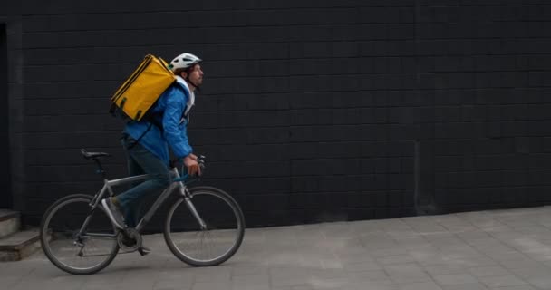 Side visning af træt mandlige kurer med madpose ridning cykel og aftørring pande på gaden. Ung mand i beskyttende hjelm leverer orden til kunden. Begrebet levering af fødevarer. – Stock-video