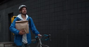 Koruyucu kasklı, yiyecek çantalı, kağıt paket taşıyan ve sokakta bisiklet süren bir erkek kurye. Müşteriye sipariş verirken gülümseyen ve yürüyen bir adam..