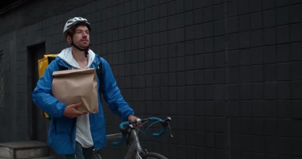 Αρσενικό courier σε προστατευτικό κράνος με τσάντα μεταφοράς τροφίμων που μεταφέρουν χάρτινο πακέτο και σπρώξιμο ποδήλατο στο δρόμο. Ο άνθρωπος χαμογελά και το περπάτημα κατά την παράδοση προκειμένου να πελάτη. — Αρχείο Βίντεο