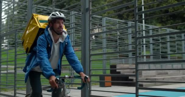 Kurier z plecakiem transportu żywności jazda na rowerze w pobliżu boiska sportowego. Człowiek w ochronnym kasku rowerowym dostarczającym zamówienie do klienta. Pojęcie usługi dostarczania żywności. — Wideo stockowe
