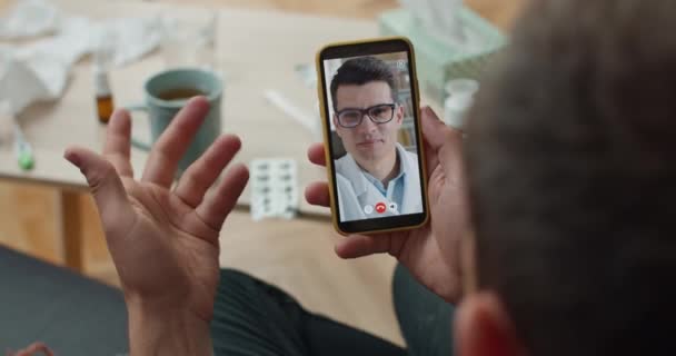 Κάτοψη του νεαρού άνδρα γιατρού συμβουλεύεται τον ασθενή με βιντεοκλήση στο smartphone και γράφοντας λεπτομέρειες. Πάνω από τον ώμο closeup οθόνη κινητής τηλεφωνίας. Έννοια της τηλεϊατρικής. — Αρχείο Βίντεο